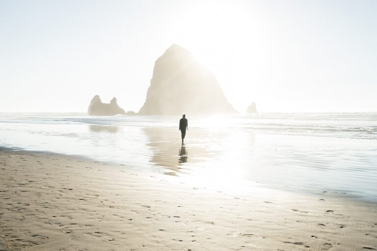Man staat stil in rimpelend water aan het strand, met zacht licht en een eiland op de achtergrond — symbool voor innerlijke balans en reflectie.